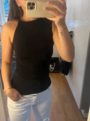 Svart tajt topp utan ärmar från H&M - Snygg kringlad detalj på ena axeln av toppen som gör den unik och cool. Perfekt till ett par vita jeans.