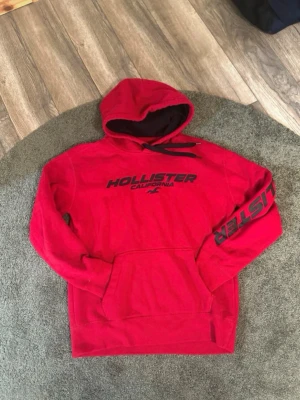 Röd hoodie från Hollister - Snygg röd hoodie från Hollister med svart tryck på bröstet och ärmen. Klassisk känguruficka framtill och justerbar huva med svarta snören. Mjuk insida och bekväm passform, perfekt för chill dagar. Materialet är bomullsblandning.