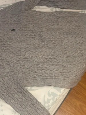 Grå kabelstickad tröja Polo Ralph Lauren - Snygg grå kabelstickad tröja från Polo Ralph Lauren med klassisk rund halsringning och diskret broderad logga på bröstet. Tillverkad i mjukt material som passar perfekt till lager på lager och ger en stilren look. 😎