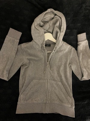 Grå hoodie från Juicy Couture - Säljer en grå zip-hoodie från Juicy Couture i mjuk bomullsblandning. Hoodien har dragkedja med JC-detalj, fickor fram och broderad logga på bröstet. Perfekt för en chill och avslappnad stil.