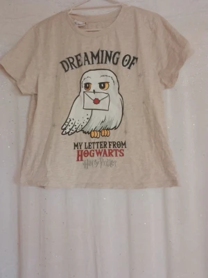 Harry Potter beige t-shirt med uggla - Beige t desert från Harry Potter RMS med tryck av Hedwig-ugglan som håller ett brev och texten 'Dreaming of my letter from Hogwarts'. T-shirten har korta sopar och sopar passform. Perfekt för alla Potterheads!