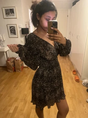 Svart och vit kortklänning med omlott - En snygg kortklänning med svart och vitt mönster, omlott-design och v-ringad hals. Klänningen har långa, puffiga ärmar och markerad midja som ger en feminin siluett. Perfekt för dig som gillar trendiga och bekväma plagg.