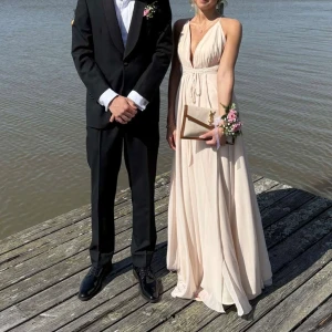 Rosa/Beige balklänning med v-ringning - Säljer nu min balklänning i ljusrosa/beige med djup v-ringning och smala axelband. Klänningen har omlottliv, markerad midja med knyt och en öppen rygg. Perfekt för bal eller sittning. 