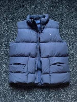 Mörkblå dunväst från Gant - Snygg mörkblå dunväst från Gant med hög krage och dragkedja framtill. Västen har två stora fickor och diskret Gant-logga med sköld på bröstet. Perfekt för lager-på-lager och håller dig varm under kyliga dagar. Nästan aldrig använd.