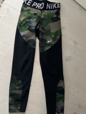 Nike Pro leggings med camomönster - Nike Pro leggings med svart bas och grönt camomönster på höft och ben. Bred resår i midjan med Nike Pro-logga. Tight passform och stretchigt material, perfekt för träning eller chill. Liten vit Nike-logga på låret.