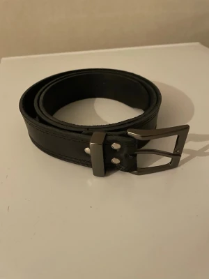 Svart klassiskt skärp i läder 118cm lång - Stilrent svart bälte i skinn med silverfärgat spänne. Perfekt accessoar för att lyfta din outfit med en tidlös och enkel design. Bältet har en klassisk form och passar till de flesta byxor.