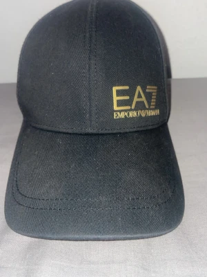 Svart keps EA7 Emporio Armani - Säljer en svart keps från EA7 Emporio Armani med guldigt tryck på framsidan. Klassisk böjd skärm och justerbar passform. Materialet är bomull och kepsen har en stilren design som passar till många outfits.