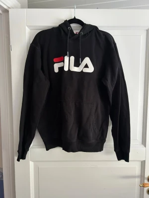 Svart hoodie från FILA med logga - Svart hoodie från FILA med stor vit och röd logga på bröstet. Klassisk modell med huva och dragsnören samt en stor magficka framtill. Perfekt för en avslappnad och sportig stil.