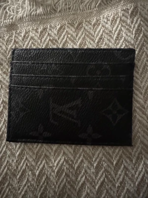 Louis Vuitton svart kortfodral - Snyggt svart kortfodral från Louis Vuitton i präglat läder med klassiskt monogrammönster. Fodralet har flera kortfack på båda sidor och en smidig, tunn design som passar perfekt i fickan. Perfekt för dig som vill ha stilren och lyxig accessoar.