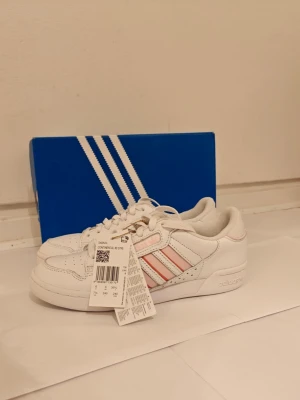 Adidas Continental 80 Stripes W - Snygga vita Adidas sneakers med klassiska tre ränder i rosa och korall på sidan. Skorna har lågt skaft, perforerad tå och Adidas-logga på hälen. Tillverkade i läder med textildetaljer och vit sula. Perfekta för en clean och sportig look. Orginalpris 1099 kr. 