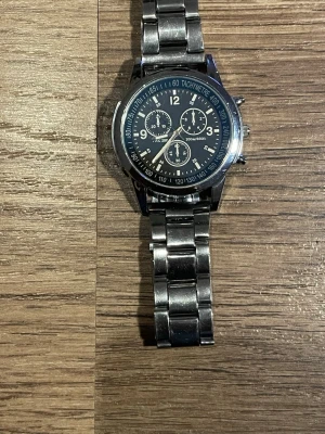 Silverfärgad kronografklocka - Stilren kronografklocka från So&Co med silverfärgad länk i metall och svart urtavla. Klockan har flera små urtavlor, tachymeter och tydliga siffror. Perfekt accessoar för dig som gillar en klassisk och sportig look.