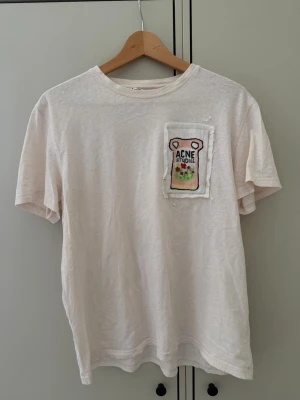 Acne Studios t-shirt med patch - Säljer en vit t-shirt från Acne Studios i storlek S. T-shirten har en unik broderad patch på bröstet med färgglada detaljer och texten 'Acne Studios'. Tillverkad i mjuk bomull och har klassisk passform med korta ärmar och rund hals. Trasig ”upphängningslapp” fotat.