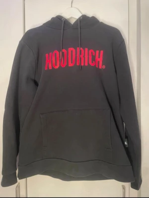 Svart HOODRICH hoodie med röd logga - Snygg svart hoodie från HOODRICH med stor röd logga på bröstet. Klassisk känguruficka framtill, justerbar huva med snören och ribbade muddar. 