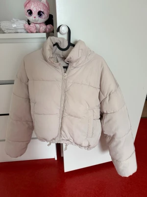 Beige pufferjacka med dragsko - Snygg beige pufferjacka med hög krage och dragkedja framtill. Jackan har två sidofickor och dragsko i nederkant för justerbar passform. Perfekt för kalla dagar och ger en trendig look.