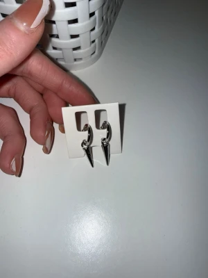Silvriga örhängen med spetsig hängande detalj - Säljer ett par silvriga örhängen med coola, spetsiga hängen. Örhängena har en minimalistisk design och är tillverkade i metall med en blank finish. Perfekta för dig som gillar edgy accessoarer och vill sticka ut lite extra.