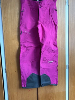 Rosa täckbyxor från Peak Performance - Säljer ett par snygga rosa täckbyxor från Peak Performance med svarta förstärkningar vid bensluten. Byxorna har flera dragkedjeförsedda fickor, justerbar midja och ventilation längs benen. Perfekta för skidåkning eller vinteraktiviteter.