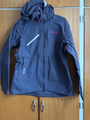 Mörklila Gore-Tex jacka  Peak Performance - Snygg mörklila vindjacka från Peak Performance med huva, dragkedja framtill och rosa detaljer. Jackan har flera fickor med dragkedja och justerbara snören vid huva och ärmslut. Perfekt för blåsiga dagar och outdoor-äventyr.
