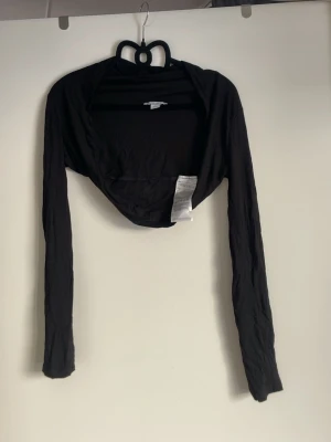 Svart bolero med långa ärmar - Snygg svart bolero med långa ärmar, perfekt att slänga över ett linne eller klänning. Tunn och mjuk i materialet, enkel att matcha till olika outfits. Kort modell som slutar under bysten och ger en stilren look.