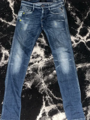 Blå slim fit jeans Replay - Säljer ett par blå slim fit jeans från Replay med slitningar och ljusa partier på låren. Jeansen har fem fickor, dragkedja och knapp, samt coola färgstänk i gult och vitt för en unik look. Klassisk denim med snygga detaljer och skön passform.