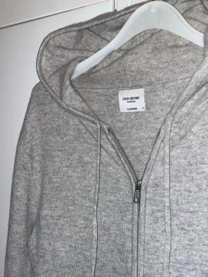 John henric kashmir Hoodie  - Mysig grå hoodie från John Henric i mjuk kashmir. Tröjan har dragkedja framtill, huva med dragsko och ribbade muddar vid ärmslut och nederkant. Perfekt för en chill och stilren look. Bra skick, använd men inga defekter. 