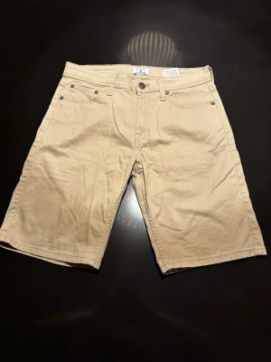 Beige shorts från Jack & Jones - Snygga beige shorts från Jack & Jones med klassisk femficksdesign och knappgylf. Tillverkade i mjuk bomull med normal passform och bälteshällor. Perfekta för varma dagar och enkel att matcha med olika stilar.