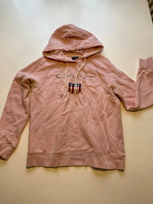 Rosa hoodie från Gant med logga - Säljer en snygg rosa hoodie från Gant med broderad logga och sköld på bröstet. Hoodien har dragsko i huvan och ribbade muddar vid ärmslut och nederkant. Perfekt för en avslappnad och trendig look.