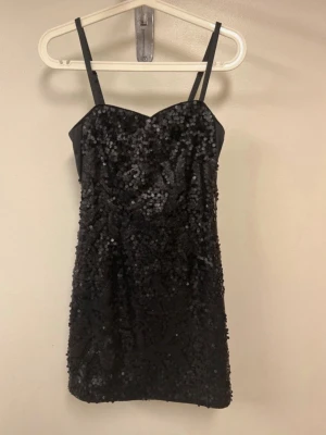 Svart paljettklänning med smala band - Snygg svart kortklänning med smala axelband och massor av glittriga paljetter över hela klänningen. Sitter figurnära och har en lätt hjärtformad urringning. Perfekt för dig som vill sticka ut och glänsa.