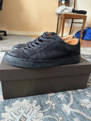 Svarta mockasneakers med låg profil - Stilrena svarta sneakers i mocka med lågt skaft och matchande svarta skosnören. Skorna har en svart gummisula och en klassisk minimalistisk design. Insidan är fodrad i ljusbrunt läder för extra komfort. Perfekta för en clean och modern look.