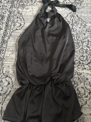 Svart halterneckklänning i satin - Snygg svart kort halterneckklänning i satinliknande material med knyt i nacken och öppen rygg. Klänningen har en lös passform och faller fint över kroppen, perfekt för dig som gillar en stilren och trendig look.