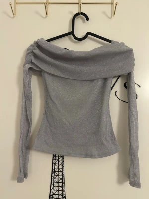 Glittrig off shoulder topp silver från NA-KD - Silverfärgad off shoulder topp med glittereffekt och långa ärmar från NA-KD i storlek XS. Toppen har en bred, draperad krage.