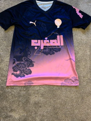 Svart och rosa fotbollströja Puma - Cool fotbollströja från Puma i svart och rosa med grafiskt mönster och arabisk text på bröstet. Tröjan har korta ärmar, V-ringning och tryckt Puma-logga. Perfekt för dig som vill sticka ut på planen.