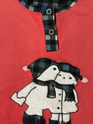 Röd fleecepyjamas med vintermotiv - Mysig röd fleecepyjamas med lång ärm och vintermotiv av två figurer med mössa och halsduk framtill. Detaljer i svart och blått rutigt tyg vid halsen och knäppning med två dekorativa knappar. Perfekt för kalla kvällar och vintermys.