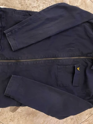 Marinblå overshirt från Lyle & Scott - Snygg marinblå overshirt från Lyle & Scott med dragkedja framtill och en bröstficka med den klassiska gula loggan. Jackan har lång ärm och är tillverkad i bomull, perfekt för lager på lager och en clean look.