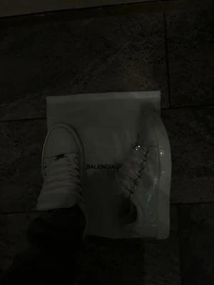 Vita Balenciaga sneakers - Säljer ett par vita sneakers från Balenciaga med klassisk låg siluett. Skorna har breda vita skosnören, metalliska detaljer och en stilren design. Tillverkade i slätt läder för en clean och exklusiv look. Perfekta för dig som gillar lyxiga streetwear-vibbar.