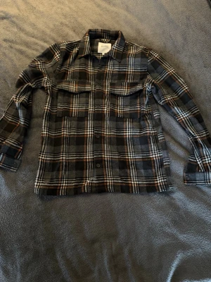 Just Junkies Overshirt - Snygg overshirt i mjuk flanell från Just Junkies med rutigt mönster i svart, grått, vitt och brunt. Två bröstfickor med lock, knäppning framtill och klassisk krage. Perfekt lager på lager-plagg för en avslappnad stil.