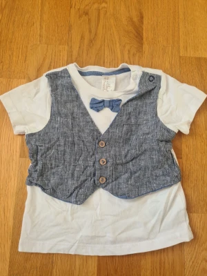 Vit t-shirt med väst och fluga H&M - Vit t-shirt från H&M med fastsydd blågrå väst och matchande fluga i storlek 68. Västen har smala ränder, dekorativa knappar och en liten ficka. Tryckknappar vid axeln för enkel påklädning. Perfekt för en dressad look!
