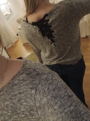 Grå tröja med snörning i ryggen - Snygg gråmelerad tröja med lång ärm och unik snörningsdetalj i svart längs ryggen. Tröjan har en avslappnad passform och är perfekt för dig som gillar trendiga detaljer. Mjuk och skön att bära.