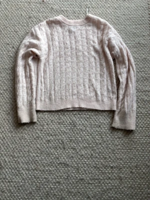 Ljusrosa kabelstickad tröja - Mysig ljusrosa kabelstickad tröja med rund hals och långa ärmar. Tröjan har ribbade muddar vid ärmslut och nederkant samt ett klassiskt kabelmönster framtill. Perfekt för kyliga dagar och enkel att matcha till olika outfits.