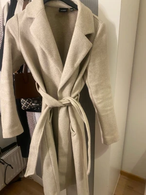 Beige kappa med knytskärp från vero Moda - Stilren beige kappa från Vero Moda med klassisk krage och knytskärp i midjan. Lång modell med mjukt tyg som ger en snygg siluett. Perfekt för dig som vill ha en enkel men trendig look till hösten eller vintern.