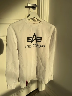 Vit sweatshirt Alpha Industries - Säljer en vit sweatshirt från Alpha Industries med stor svart logga och text på bröstet. Tröjan har rund halsringning, långa ärmar och ribbade muddar vid ärmslut och nederkant. Perfekt för en clean och avslappnad stil.