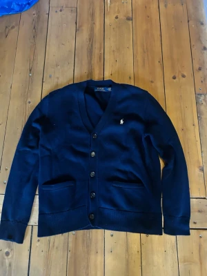 Mörkblå cardigan från Polo Ralph Lauren - Klassisk mörkblå cardigan från Polo Ralph Lauren med v-ringning och knäppning framtill. Tröjan har två fickor, ribbade muddar och den ikoniska vita logotypen broderad på bröstet. Perfekt för en stilren och avslappnad look. Priset är ej hugget i sten 