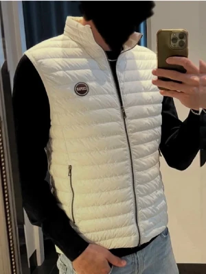 Vit dunväst från Colmar - Snygg vit dunväst från Colmar med quiltad design och hög krage. Västen har två dragkedjeförsedda fickor och en Colmar-logga på bröstet. Perfekt att slänga över en hoodie eller tröja för en clean och sportig look.