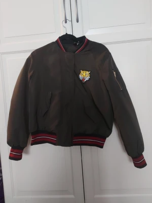 Svart bomberjacka med tigerpatch - Cool svart bomberjacka med broderad tigerpatch på bröstet. Jackan har röda och vita ränder på muddar och krage, dragkedja framtill och ficka med dragkedja på ärmen. Perfekt för dig som vill sticka ut med en unik detalj.