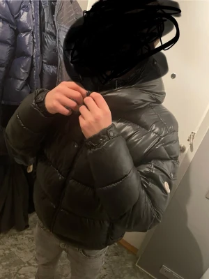 Svart pufferjacka med huva - Säljer en svart moncler pufferjacka med stor huva och quiltad design. Jackan har hög krage, dragkedja framtill och en patch på ärmen. Perfekt för kalla dagar och riktigt skön att bära. Passar dig som gillar en stilren. Häng saken är sönder men det syns inte. Det är lätt att laga. Den passar folk mellan 170cm till 185cm
