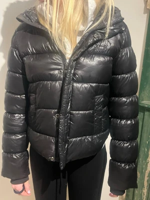 Svart pufferjacka med huva - Snygg svart pufferjacka med glansig finish och stor huva. Jackan har breda kanaler, dragkedja och tryckknappar framtill samt två fickor på sidorna. Perfekt för kalla dagar och ger en cool streetstil.har en liten lagning längst ner vid blixtlåset insidan, syns inte när jackan är knäppt. Vill ni ha mer bilder så skickar jag. 