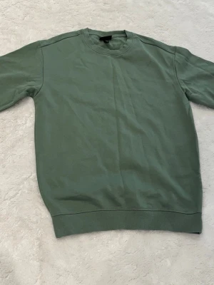 Grön sweatshirt utan huva - En enkel och stilren grön sweatshirt i mjukt bomullsmaterial. Tröjan har rund halsringning, långa ärmar och ribbade muddar vid ärmslut och nederkant. Perfekt för en avslappnad look och lätt att matcha med jeans eller joggers.