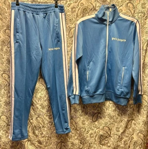 Blått tracksuit set från Palm Angels - Snyggt blått tracksuit set från Palm Angels med vita ränder längs ärmar och ben. Setet består av en zip-jacka med hög krage och två fickor samt matchande byxor med resår i midjan. Materialet är mjukt och stretchigt, perfekt för en sportig och avslappnad look.