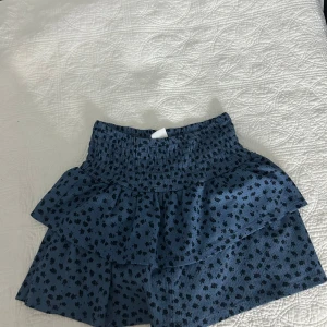 Blå blommig volangkjol från Zara - Supersöt blå kortkjol från Zara med svarta små blommor och volangdetaljer. Kjolen har en smockad midja som gör den bekväm och sitter snyggt. Materialet är mjukt och har en lätt struktur, perfekt för dig som gillar en chill och trendig stil.