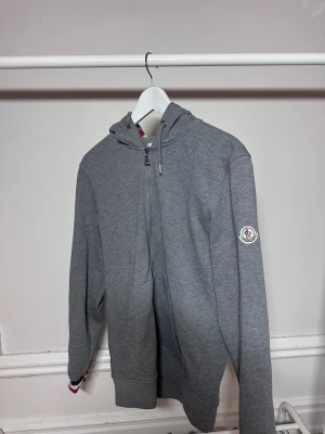 Grå Moncler hoodie  - Snygg grå hoodie från Moncler med dragkedja framtill och klassisk huva. Tröjan har ribbade muddar och Moncler-logga på ärmen. Tillverkad i mjukt bomullsmaterial som känns skönt mot huden. Perfekt för en avslappnad och stilren look.Perfekt för dig som vill ha ett plagg som kombinerar komfort, kvalitet och exklusivt varumärke. Nypris betydligt högre. 📦 Kan skickas snabbt 💬 Hör av dig vid frågor eller om du vill ha fler bilder