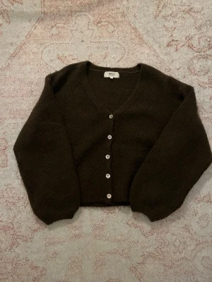 Mörkbrun kofta från Gina Tricot - Mysig mörkbrun kofta från Gina Tricot med v-ringning och fem vita knappar framtill. Koftan har ballongärmar och ribbade muddar vid ärmslut och nederkant. Perfekt för lager på lager och enkel att styla till olika outfits.